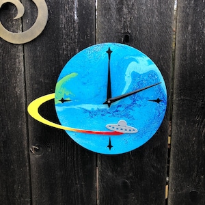 May include: A round, blue clock with a planet design, featuring a yellow ring and a red streak. A silver UFO is depicted on the clock face. Black clock hands and markers complete the design. The clock is mounted on a dark wooden fence.