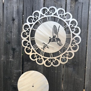 May include: A metal wall clock with a hummingbird design and a decorative scrollwork border. The clock face has Roman numerals and the hummingbird is in silhouette. The clock is mounted on a wooden fence.