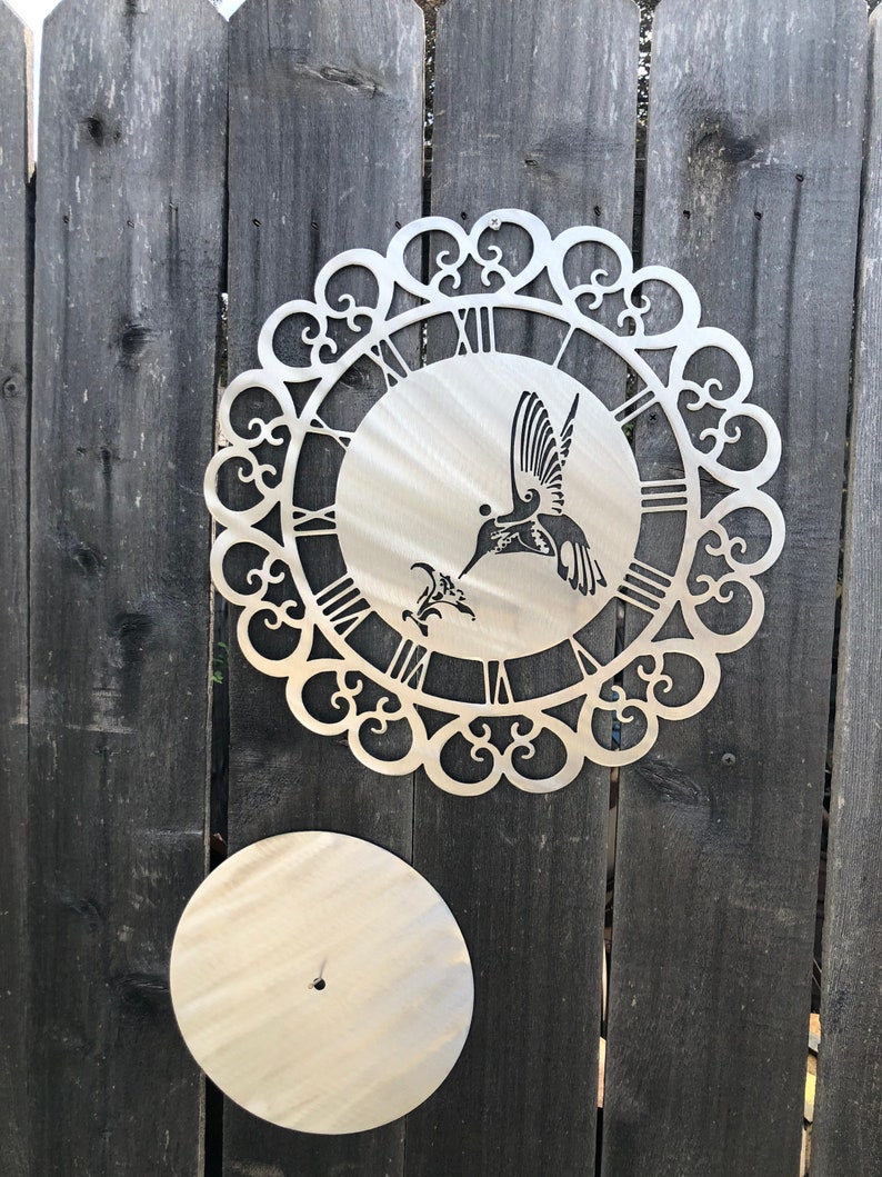 May include: A metal wall clock with a hummingbird design and a decorative scrollwork border. The clock face has Roman numerals and the hummingbird is in silhouette. The clock is mounted on a wooden fence.