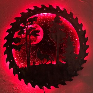 Op de afbeelding: Een rood verlicht metalen zaagblad met een silhouet van een wolf die huilt naar de maan in een bos sc&egrave;ne.