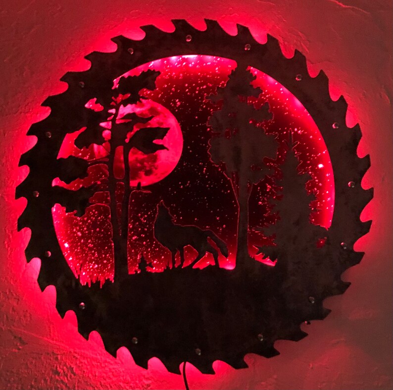 Op de afbeelding: Een rood verlicht metalen zaagblad met een silhouet van een wolf die huilt naar de maan in een bos sc&egrave;ne.
