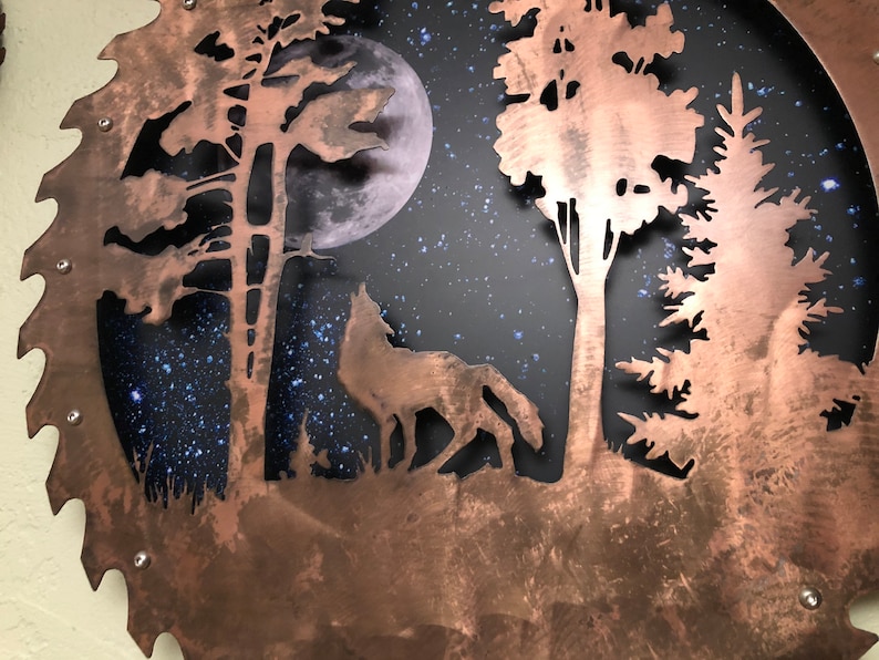 Op de afbeelding: Een koperkleurig metalen zaagblad met een uitgesneden ontwerp van een wolf die huilt naar de maan in een bossetting. De achtergrond is donkerblauw met witte sterren.