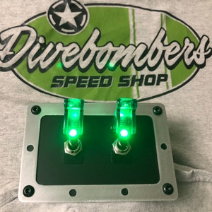 May include: Two green illuminated toggle switches mounted in a silver metal panel with black trim. The panel has six screw holes and is labeled "Divebombers Speed Shop".