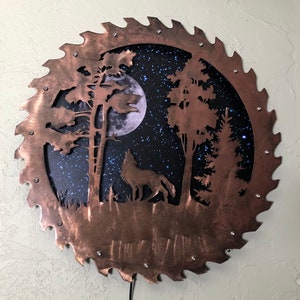 Op de afbeelding: Een cirkelvormig wandkunstwerk van koper met een zaagbladontwerp. Het midden van het stuk toont een silhouet van een wolf die huilt naar de maan in een bos sc&egrave;ne. De achtergrond is donkerblauw met witte sterren.