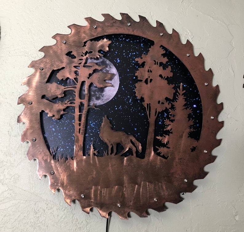 Op de afbeelding: Een cirkelvormig wandkunstwerk van koper met een zaagbladontwerp. Het midden van het stuk toont een silhouet van een wolf die huilt naar de maan in een bos sc&egrave;ne. De achtergrond is donkerblauw met witte sterren.