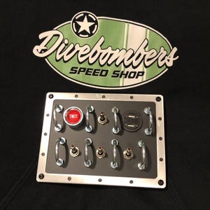 May include: A black panel with a silver frame featuring various switches, a red button, and silver handles. The text "Divebombers SPEED SHOP" is above the panel in a green and white oval.