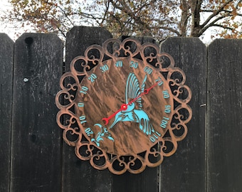 Op de afbeelding: Een koperkleurige metalen thermometer met een decoratief kolibrie-ontwerp. De thermometer heeft een zwarte achtergrond en witte cijfers. De kolibrie is blauw en rood.