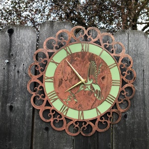 Könnte beinhalten: Eine kupferfarbene Metalluhr mit grünem Zifferblatt und einem Kolibri-Design. Die Uhr ist an einem Holzzaun befestigt.