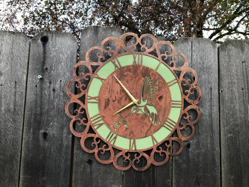 May include: A copper-colored metal clock with a green face and a hummingbird design. The clock is mounted on a wooden fence.