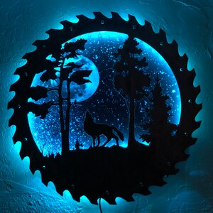 Op de afbeelding: Een blauw verlicht wandkunstwerk dat een wolf voorstelt die huilt naar de maan in een bossetting. De kunst is gemaakt van metaal en heeft een zaagbladontwerp rond de rand.