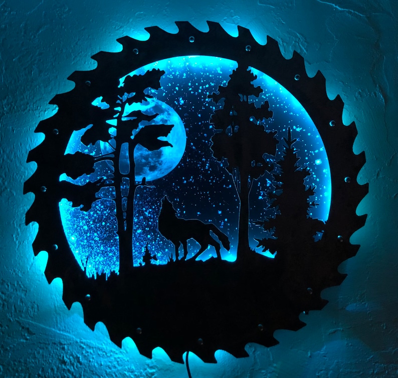 Op de afbeelding: Een blauw verlicht wandkunstwerk dat een wolf voorstelt die huilt naar de maan in een bossetting. De kunst is gemaakt van metaal en heeft een zaagbladontwerp rond de rand.