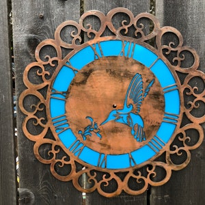 May include: A copper-colored metal clock with a blue face and a hummingbird design. The clock is surrounded by a decorative metal frame.