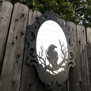 May include: A black metal mirror with an ornate frame and a raven silhouette in the center. The mirror is hanging on a wooden fence.