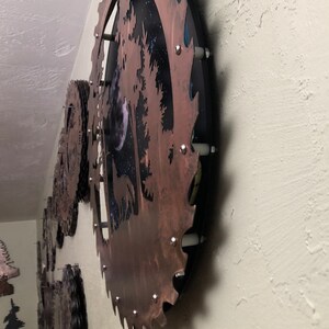 Op de afbeelding: Een cirkelvormig metalen wandkunstwerk met een rustieke bruine afwerking. De kunst toont een silhouet van een boom met een volle maan en sterren op de achtergrond. Het metaal is getextureerd en heeft een verweerde uitstraling.