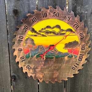 May include: A circular metal thermometer with a sunset scene and a red needle. The thermometer is marked with numbers from 0 to 110.