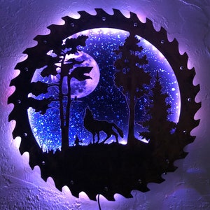 Op de afbeelding: Een zwart metalen silhouet van een wolf die huilt naar de maan in een bossetting. Het silhouet is uitgesneden uit een cirkelvormige zaagblad met een blauw LED-licht dat door de achterkant schijnt. De maan en de sterren zijn ook uit het metaal uitgesneden.