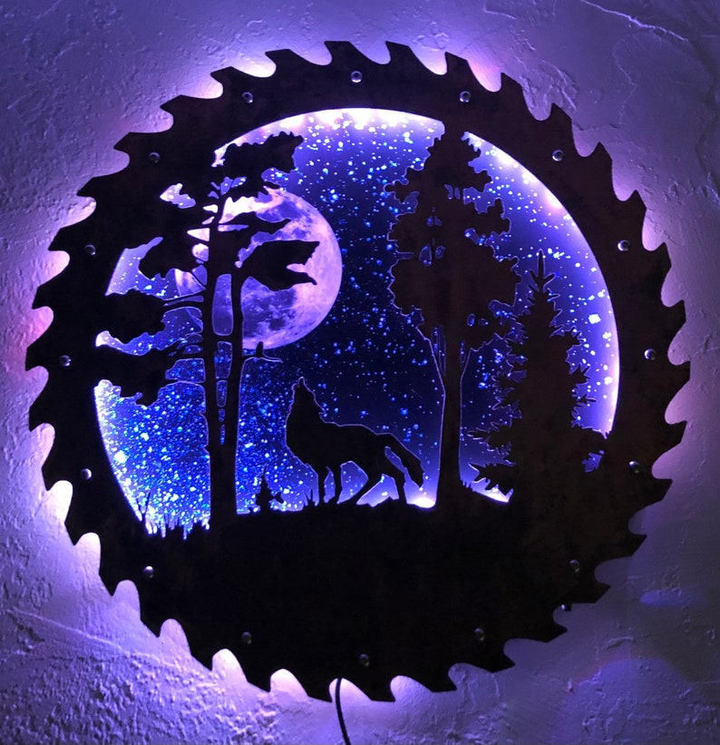 Op de afbeelding: Een zwart metalen silhouet van een wolf die huilt naar de maan in een bossetting. Het silhouet is uitgesneden uit een cirkelvormige zaagblad met een blauw LED-licht dat door de achterkant schijnt. De maan en de sterren zijn ook uit het metaal uitgesneden.