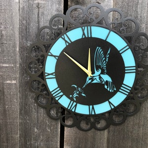 May include: A black metal wall clock with a blue and black face. The clock features a hummingbird design and Roman numeral hour markers.