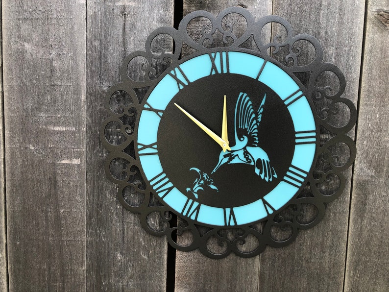 May include: A black metal wall clock with a blue and black face. The clock features a hummingbird design and Roman numeral hour markers.