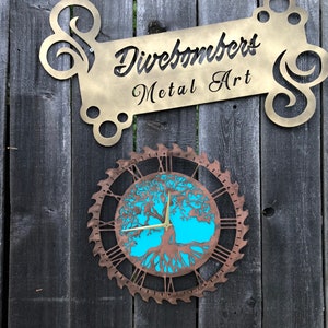 May include: A metal art sign and clock are mounted on a wooden fence. The sign reads "Divebombers Metal Art" in a gold color. The clock has a copper-colored saw blade frame with a tree of life design and Roman numerals. The clock face is turquoise.