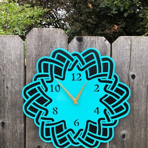 Puede incluir: Un reloj de pared turquesa con un diseño de nudo celta negro alrededor de la esfera. El reloj tiene manecillas doradas y números negros. El reloj está montado en una valla de madera desgastada.