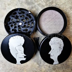 May include: A black metal herb grinder featuring a silver graphic of Beavis and Butt-Head on the lids. The set includes a top lid with sharp teeth, a middle section with a fine mesh screen, and a bottom collection chamber, all in a circular shape.