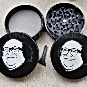 Puede incluir: Un molinillo de hierbas negro de cuatro piezas con una ilustración gráfica blanca de un hombre con gafas y pelo rizado en la tapa. El conjunto incluye un compartimento con malla, una cámara de trituración con dientes y una pequeña espátula.