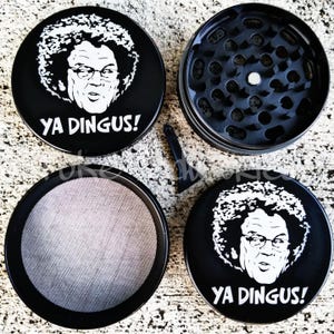 May include: A black four-piece herb grinder featuring a white graphic print of a person with an afro and glasses, accompanied by the text YA DINGUS! The set includes a lid, a grinding chamber with metal teeth, a mesh screen, and a small black scraper.