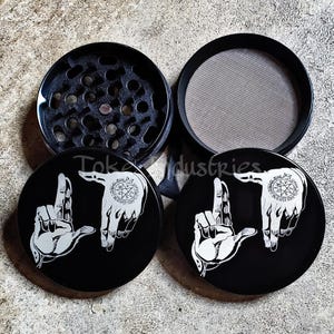 May include: A black circular herb grinder with four components, featuring a white graphic of two hands forming an L-shape with a pentagram symbol on the palm. The interior shows a grinding chamber with teeth and a fine mesh screen filter.