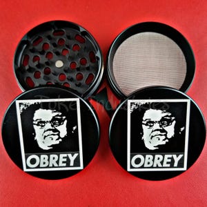 May include: A black circular herb grinder consisting of four pieces. The top lids feature a white graphic print of a person's face with the text OBREY below. The interior components include a metal mesh screen and a grinding chamber with sharp teeth.