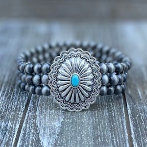 Puede incluir: Un brazalete de plata con una piedra turquesa en el centro de un concho de plata. El brazalete está hecho de cuentas grises.