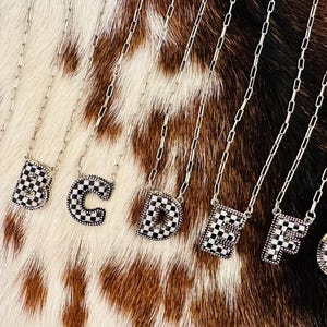 Puede incluir: Siete collares de plata con colgantes de letras a cuadros en blanco y negro. Las letras deletrean "ABCDEFG".