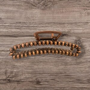 May include: A copper-colored hair claw clip. The clip has a spring mechanism and a series of small, rounded beads along the curved arms. The clip is designed to hold hair in place and is made of metal.