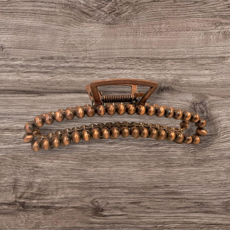 May include: A copper-colored hair claw clip. The clip has a spring mechanism and a series of small, rounded beads along the curved arms. The clip is designed to hold hair in place and is made of metal.