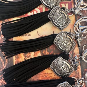 May include: Close-up of several keychains with black tassels. Each keychain features a silver-colored, ornate buckle with a different letter. The background includes a vintage-style print with the word "WEST".