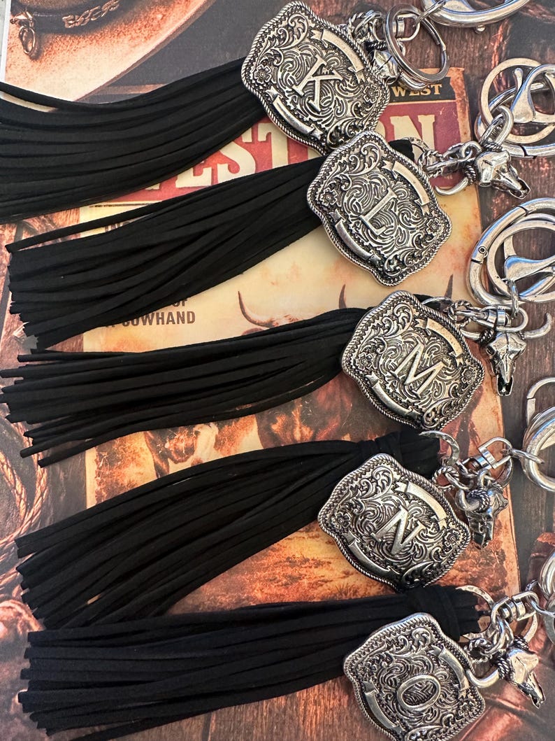 May include: Close-up of several keychains with black tassels. Each keychain features a silver-colored, ornate buckle with a different letter. The background includes a vintage-style print with the word "WEST".