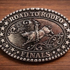 May include: Oval silver-colored belt buckle with the words "ROAD TO RODEO FINALS". The buckle features a rodeo scene with a rider on a bucking bull, surrounded by stars and decorative flourishes.