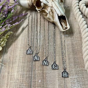 May include: Five silver necklaces with letter charms in the shape of a cattle brand. The charms spell out the word "RANCH".