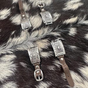 May include: Three silver watch bands with intricate geometric designs and brown leather straps. The bands are laid out on a brown and white cowhide surface.