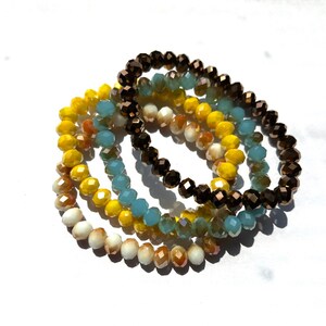 May include: Four beaded bracelets in brown, yellow, blue, and white. The bracelets are stacked on top of each other.