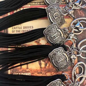 May include: Close-up of several keychains with silver-colored, engraved western-style belt buckle charms. Each keychain has a black tassel and a small bull skull charm. The background features text and images.