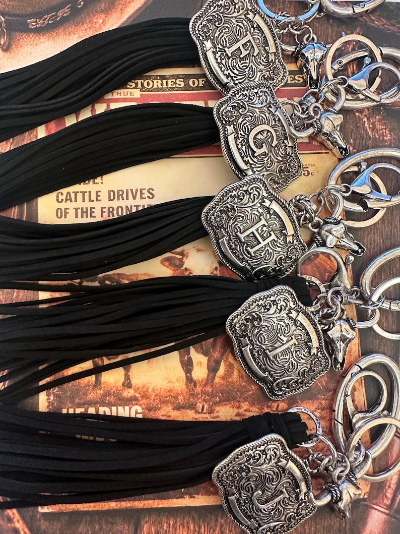 May include: Close-up of several keychains with silver-colored, engraved western-style belt buckle charms. Each keychain has a black tassel and a small bull skull charm. The background features text and images.