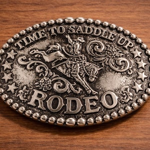 May include: Oval silver-colored belt buckle with the words "TIME TO SADDLE UP" and "RODEO" in raised lettering. The buckle features a cowboy riding a bucking bronco, surrounded by stars and decorative scrollwork.