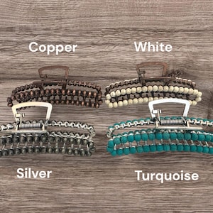 May include: Four beaded hair claw clips in copper, white, silver, and turquoise. Each clip has a metal clasp and is adorned with rows of beads. The clips are displayed on a wooden surface, with the color names written below each clip.