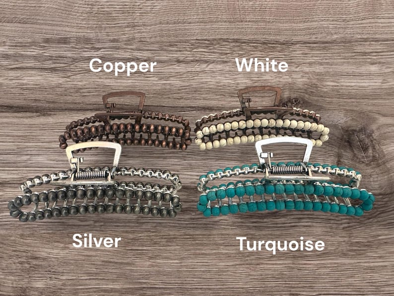 May include: Four beaded hair claw clips in copper, white, silver, and turquoise. Each clip has a metal clasp and is adorned with rows of beads. The clips are displayed on a wooden surface, with the color names written below each clip.