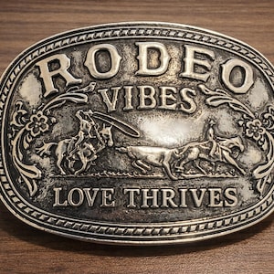 May include: Oval silver-colored belt buckle with the words "RODEO VIBES LOVE THRIVES" in raised lettering. The buckle features detailed illustrations of rodeo riders and floral accents. The buckle is set against a wood grain background.