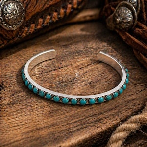 May include: Silver cuff bracelet with turquoise stones. The open-ended bracelet features a row of small, round turquoise stones set in a silver band. The bracelet is displayed on a wooden surface, with leather and metal accents in the background.