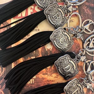 May include: Silver-toned initial keychains with black tassels. Each keychain features a decorative, engraved letter A, B, C, D, or E. The keychains have a western-style design and are attached to a silver keyring.