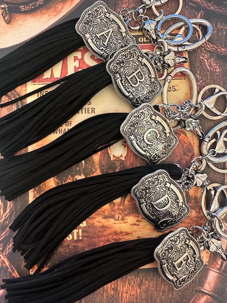 May include: Silver-toned initial keychains with black tassels. Each keychain features a decorative, engraved letter A, B, C, D, or E. The keychains have a western-style design and are attached to a silver keyring.