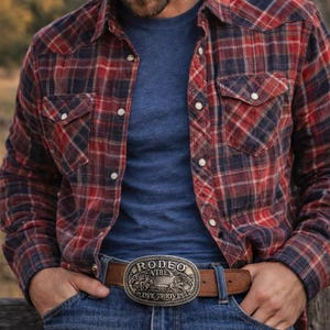 May include: A person wearing a red and navy plaid button-down shirt over a blue t-shirt, blue jeans, and a cowboy hat. A brown leather belt with a silver rodeo buckle that reads "Rodeo Vibes Love Thrives" is visible.